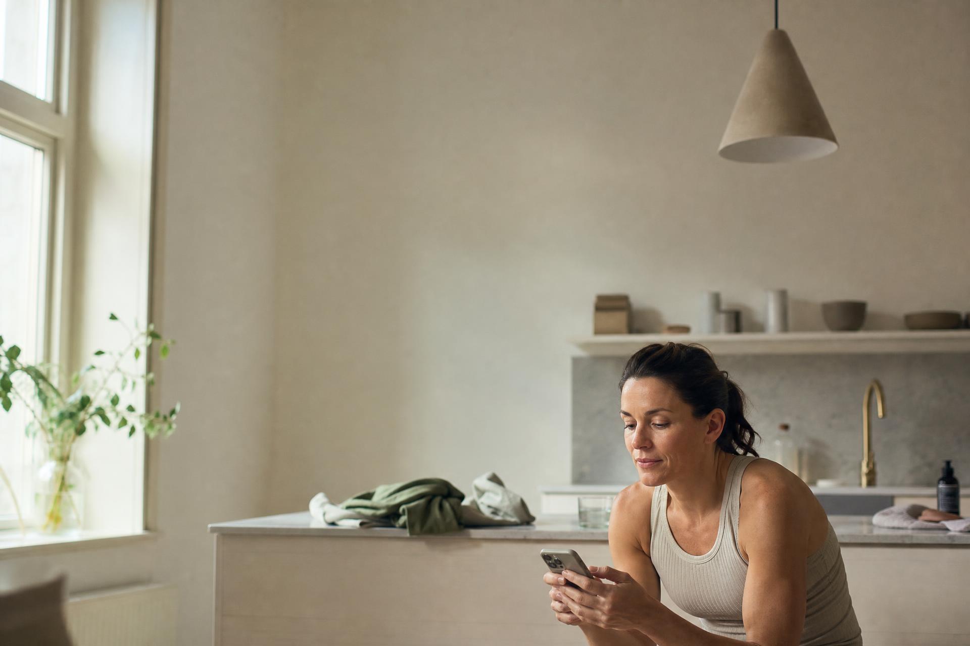 Active woman in her thirties reading a peptide consultation on her phone in a sunlit kitchen